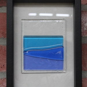 May include: A framed piece of abstract art featuring layers of blue glass. The glass is layered to create a wave-like effect. The frame is black and the background is white.