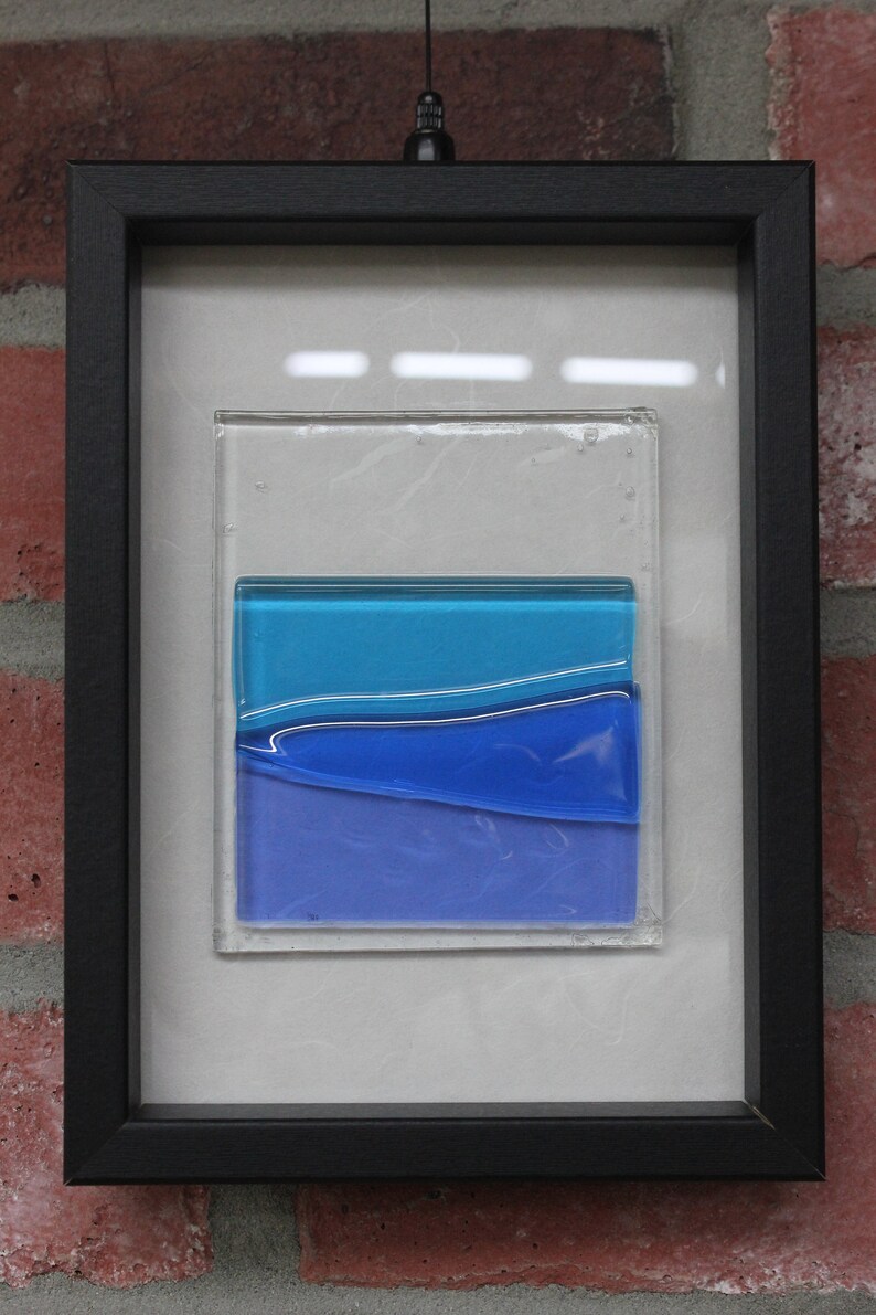 May include: A framed piece of abstract art featuring layers of blue glass. The glass is layered to create a wave-like effect. The frame is black and the background is white.