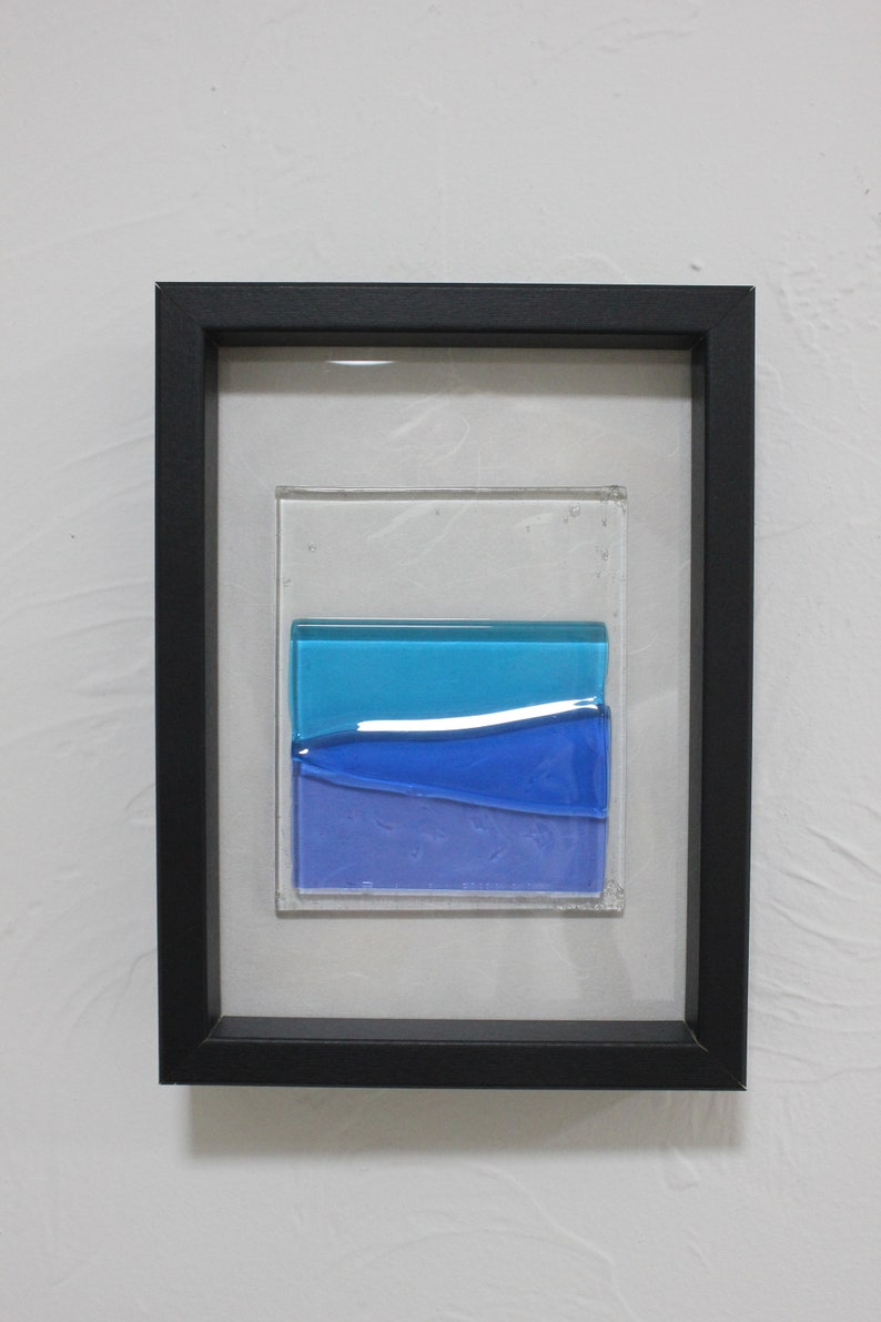 May include: A framed piece of art featuring a layered glass design with blue and purple hues. The glass is set in a black frame.