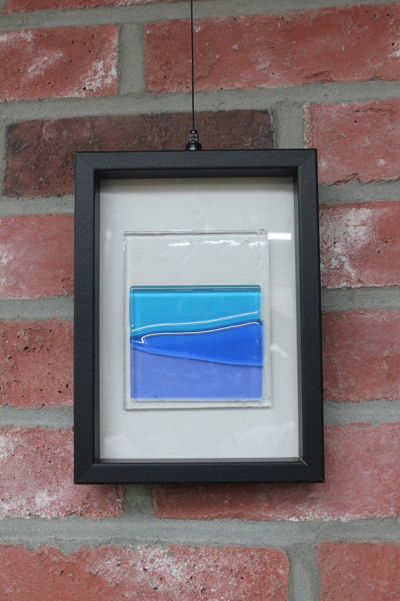 May include: A framed piece of abstract art featuring layered blue glass panels. The glass panels are set against a white background and framed in black.