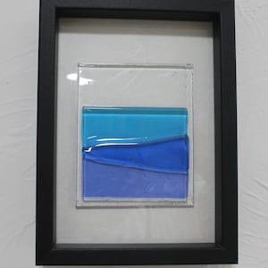 May include: A framed piece of art featuring a layered glass design with blue and purple hues. The glass is set in a black frame.