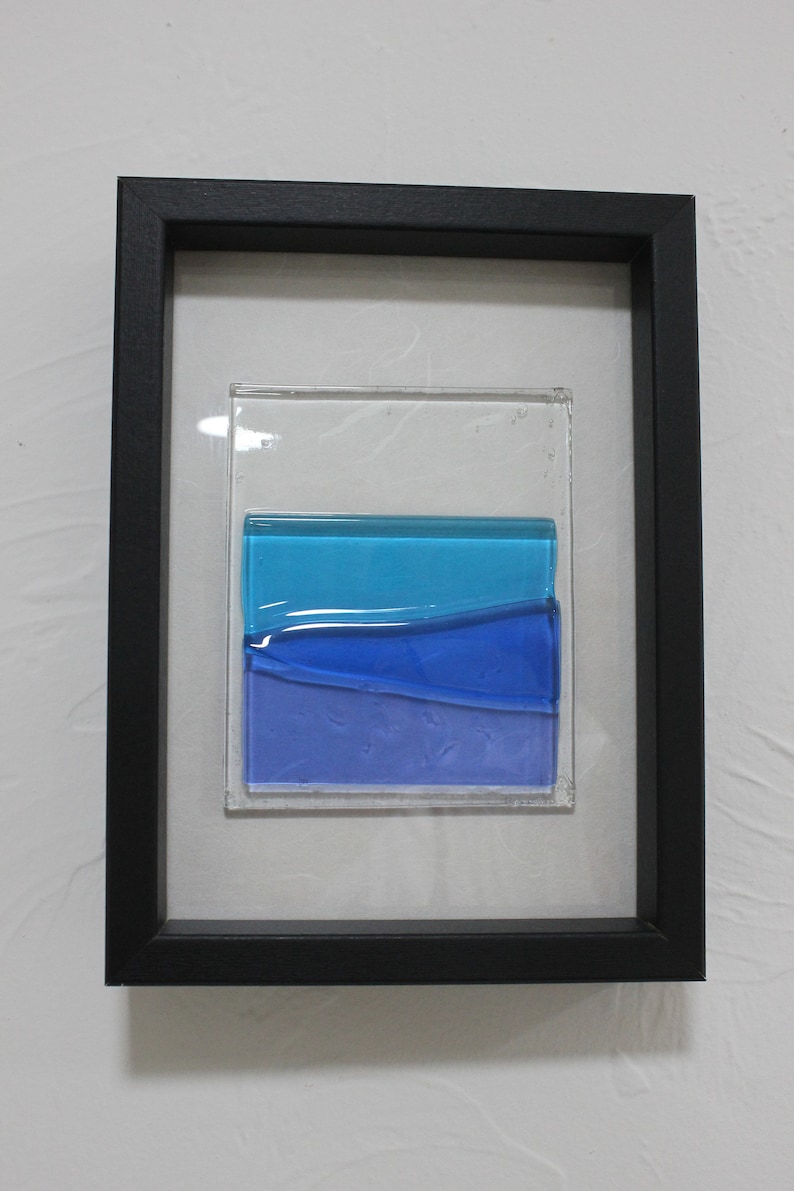 May include: A framed piece of art featuring a layered glass design with blue and purple hues. The glass is set in a black frame.