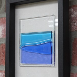 May include: A framed abstract art piece featuring layered blue glass panels. The glass panels are arranged in a wave-like pattern, creating a sense of movement and depth. The piece is framed in a black frame.