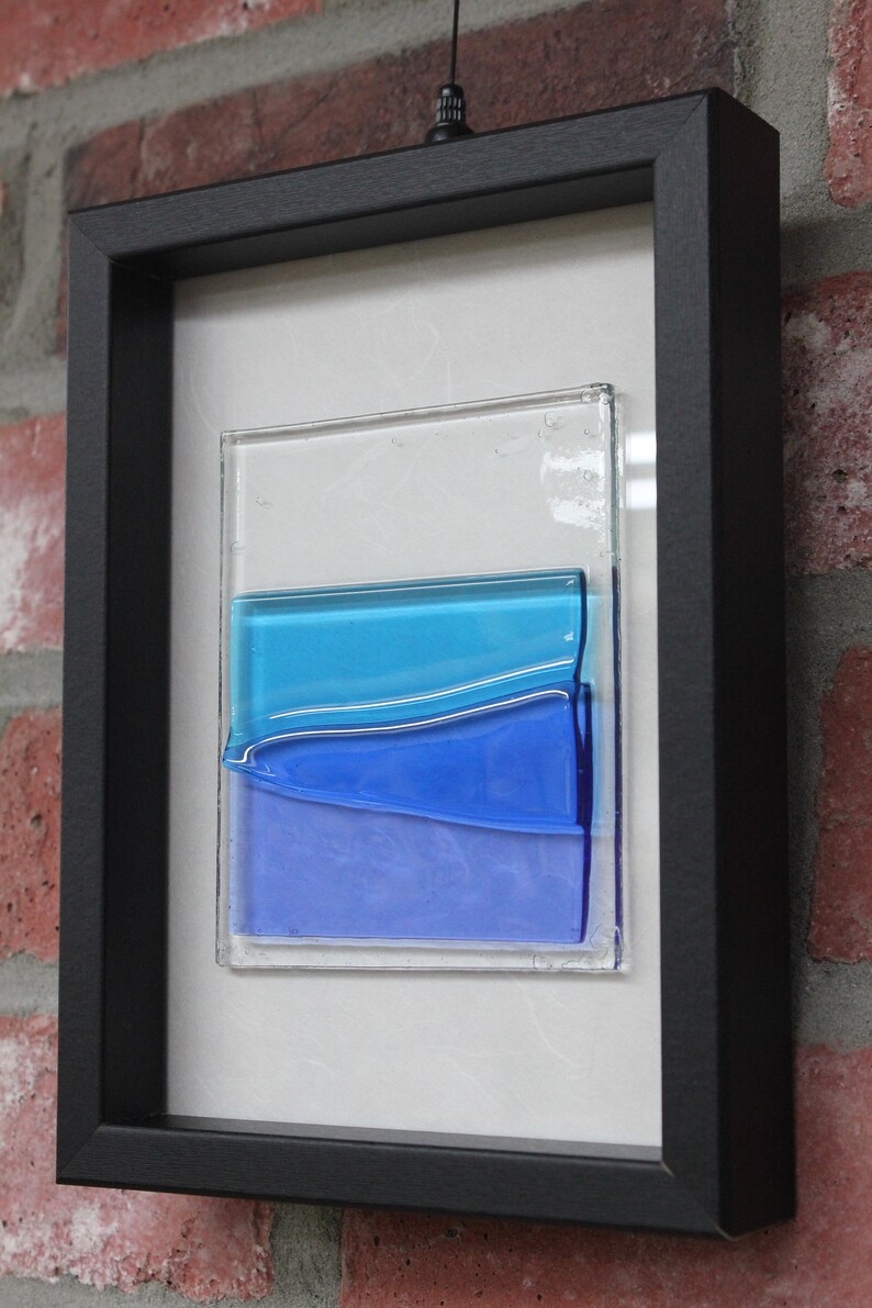 May include: A framed abstract art piece featuring layered blue glass panels. The glass panels are arranged in a wave-like pattern, creating a sense of movement and depth. The piece is framed in a black frame.