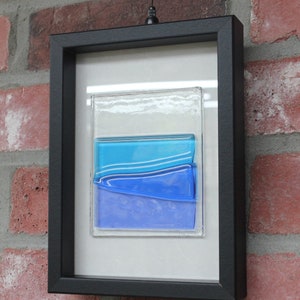 May include: A framed piece of art featuring layered blue glass with a white background. The frame is black and the glass is textured.