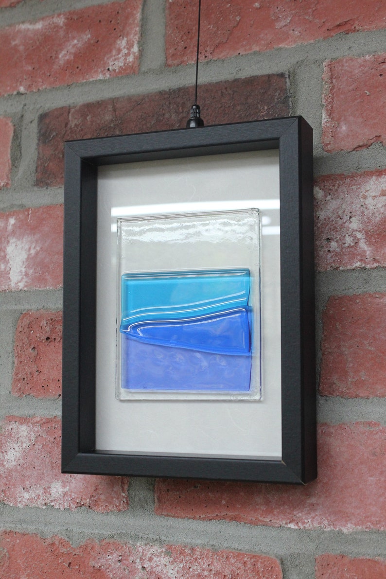 May include: A framed piece of art featuring layered blue glass with a white background. The frame is black and the glass is textured.