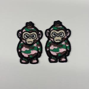 Può includere: Due toppe ricamate raffiguranti un personaggio scimmia stilizzato. Ogni scimmia presenta un motivo mimetico nei toni del verde, rosa e marrone, delineato da un bordo nero spesso. I volti sono beige chiaro con dettagliate cuciture nere.