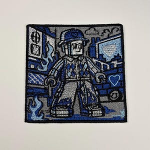 Könnte beinhalten: Ein quadratischer Stickaufnäher mit einer stilisierten Blockfigur, die eine Kappe und eine Jacke mit Rautenmuster trägt. Das Design verwendet Blau-, Grau- und Schwarztöne und enthält Flammenmotive, eine Herzform und einen Ziegelhintergrund.