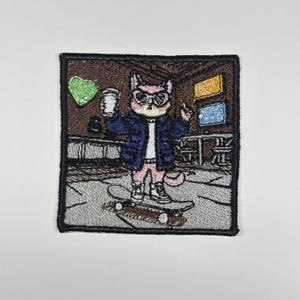 May include: A square embroidered patch featuring a cat wearing sunglasses, a dark blue jacket, and light pink pants while riding a skateboard. The cat holds a white cup in one paw. The background depicts a brown room with a green heart shape.