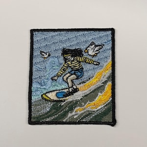 May include: A rectangular embroidered patch featuring a person with dark hair wearing a yellow and white striped long-sleeve shirt and blue shorts, surfing on a surfboard over a wave with yellow foam, accompanied by two white seagulls in the sky.