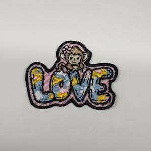 May include: An embroidered patch featuring the word LOVE in a bubbly font with a tie-dye pattern of blue, yellow, and pink. A small brown monkey with a pink flower on its head sits atop the letter O. The entire design has a thick black border.