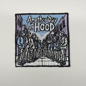 May include: A square embroidered patch featuring a street scene with a group of people standing on both sides of a path. The text Another Day in the HOOD is stitched in the centre. The colour palette includes shades of light blue, purple, and black.