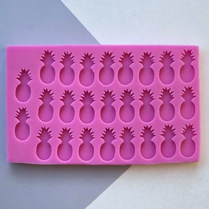 Op de afbeelding: Een rechthoekige, felroze siliconen mal met 24 ananasvormen. De mal is ontworpen voor het maken van kleine, decoratieve ananasvormige items. De achtergrond is een mix van grijs en wit.