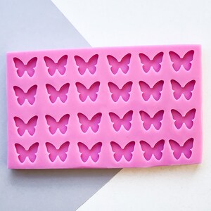 Op de afbeelding: Een roze siliconen mal met 24 vlindervormige holtes. De rechthoekige mal is ontworpen voor het maken van decoratieve elementen. De vlinders zijn gelijkmatig verdeeld en gedetailleerd, perfect voor knutsel- of bakprojecten.