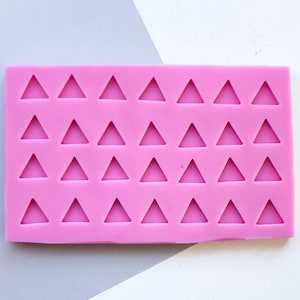 Peut inclure: Un moule en silicone rose vif de forme rectangulaire. Il comporte 30 cavités triangulaires disposées en grille. Le fond est un mélange de blanc et de gris.