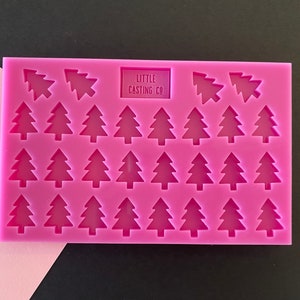 May include: A vibrant pink silicone mould with a rectangular form, showcasing numerous miniature Christmas tree designs. The text "LITTLE CASTING CO" is printed on the mould. The background is a mix of pink and black.