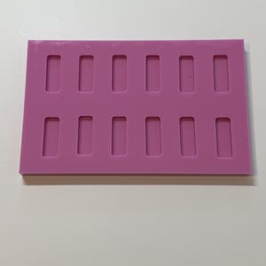 May include: A rectangular pink silicone mould featuring twelve identical recessed cavities arranged in two horizontal rows of six. Each cavity is a rounded rectangle shape, designed for casting or moulding small items in a uniform format.