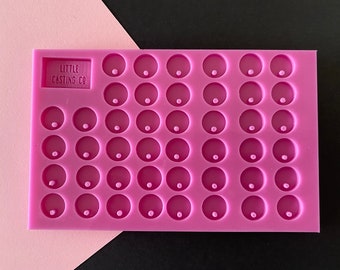 May include: A pink silicone mold with multiple circular cavities, each with a small central indentation. The mold has the text "LITTLE CASTING CO" in a rectangular area. The background is a split of pink and black.
