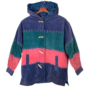 May include: A color-blocked jacket with a hood, featuring sections of blue, green, and red suede-like material. The jacket has wooden toggle closures and decorative stitching. The sleeves are long and the jacket is hanging on a wooden hanger.