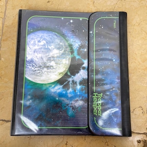 May include: A black Trapper Keeper binder with a space-themed design. The cover shows a planet with blue, green, and white colours, set against a starry background. The binder is open, revealing the interior.