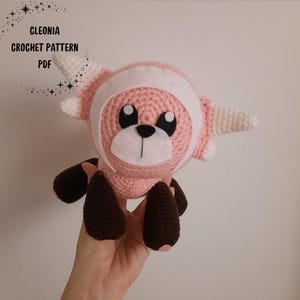 May include: A handmade pink crochet plush toy with a white and pink head covering and dark brown feet. The toy has large black eyes and a white muzzle. The text "CLEONIA CROCHET PATTERN PDF" is in the upper left corner.