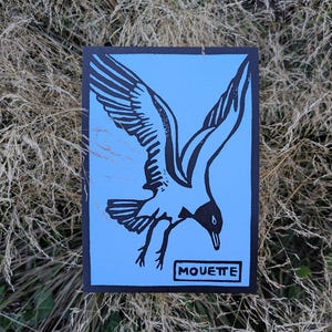 Può includere: Una stampa artistica rettangolare con un'illustrazione in bianco e nero di un gabbiano in volo su sfondo azzurro. La parola "MOUETTE" è in un riquadro bianco in basso. La stampa ha un bordo nero.