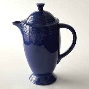 May include: A deep cobalt blue ceramic coffee pot with a glossy finish. The vessel features a tall, tapered body, a curved handle, a pointed spout, and a matching lid with a small knob handle. The base and lid are decorated with concentric rings.