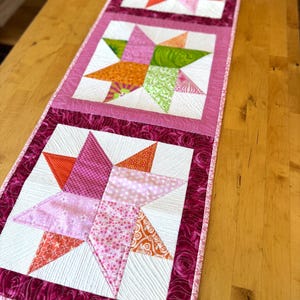 May include: A quilted table runner featuring three star blocks in shades of pink, orange, and green. Each star is set against a white background and framed with a dark pink border. The runner is placed on a wooden surface.