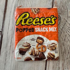May include: Orange zippered pouch featuring the Reese's brand name and "POPPED SNACK MIX" text. The pouch design includes images of Reese's Peanut Butter Cup Minis, pretzels, and chocolate drizzled popcorn.