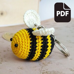 May include: A crocheted bee keyring with black and yellow stripes, white wings, and black eyes. The bee is attached to a silver key ring.