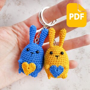 May include: Two crocheted bunny keychains, one blue and one yellow, each holding a heart-shaped keychain. The keychains are attached to a silver key ring.
