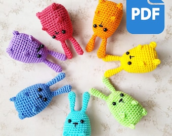 May include: A set of seven crocheted bunny toys in rainbow colors. The bunnies are arranged in a circle on a white background. The bunnies have black eyes and a black nose. The bunnies are all about 4 inches tall.
