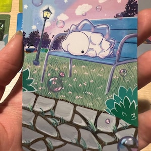 May include: A card with an illustration of a white creature on a blue park bench. The background depicts a park scene with a lamppost, bubbles, and a pink and blue sky. The card is held in a hand.