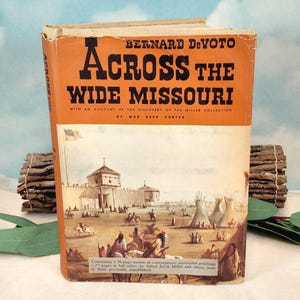 May include: A hardcover book titled Across the Wide Missouri by Bernard DeVoto. The orange dust jacket features black text and a historical illustration of a fort, teepees, and figures on horseback. The book includes an account of the Miller collection.