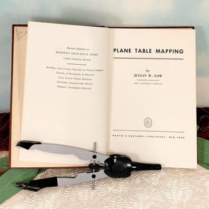 May include: An open book titled "Plane Table Mapping" by Julian W. Low, with a compass on top. The book is part of the Harper's Geoscience Series. The compass is silver and black.