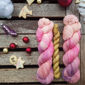 Puede incluir: Dos ovillos de hilo, uno rosa y morado degradado y otro dorado, se muestran sobre una superficie de madera con adornos navideños y galletas.