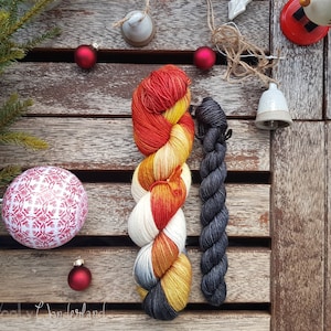 Puede incluir: Dos ovillos de hilo, uno con un degradado de color rojo, naranja y amarillo y el otro gris oscuro. El hilo está dispuesto sobre una superficie de madera con adornos rojos y blancos.