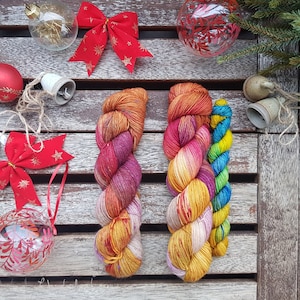 Puede incluir: Tres ovillos de hilo en varios colores, incluyendo rojo, naranja, amarillo y azul. El hilo está dispuesto sobre una superficie de madera con decoraciones navideñas.