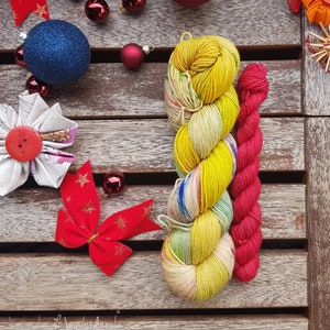 Puede incluir: Dos ovillos de hilo, uno amarillo y otro rojo, sobre una superficie de madera con decoraciones navideñas. El hilo amarillo tiene un patrón multicolor y el hilo rojo es liso.