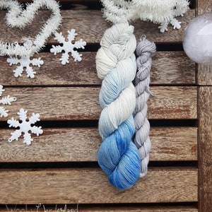 Puede incluir: Dos ovillos de hilo, uno azul y blanco degradado y otro gris, se muestran sobre una superficie de madera con decoraciones de copos de nieve blancos.