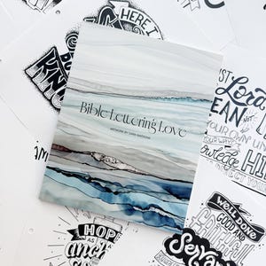 Pode incluir: Um livro intitulado "Bible Lettering Love" com um design abstrato em aquarela em tons de azul, cinza e marrom. O livro &eacute; cercado por v&aacute;rias p&aacute;ginas brancas com cita&ccedil;&otilde;es e frases inspiradoras manuscritas em preto.