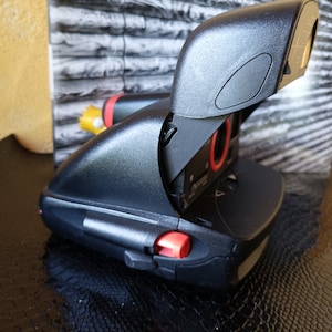 Peut inclure: Un appareil photo instantané noir avec un bouton rouge et un flash noir. L'appareil photo est ouvert et l'objectif est visible.