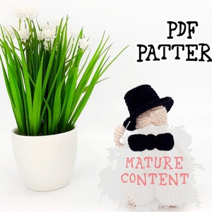Könnte beinhalten: Eine gehäkelte Amigurumi-Puppe mit einem schwarzen Zylinder und einer Fliege. Die Puppe hält ein Schild mit der Aufschrift "Mature Content". Die Puppe sitzt auf einer weißen Oberfläche vor einer Topfpflanze mit weißen Blüten.