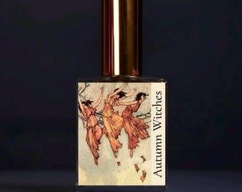 May include: A clear glass perfume bottle with a metallic gold cap. The rectangular label features an illustration of three figures in flowing orange dresses dancing among branches, with the text Autumn Witches printed vertically.