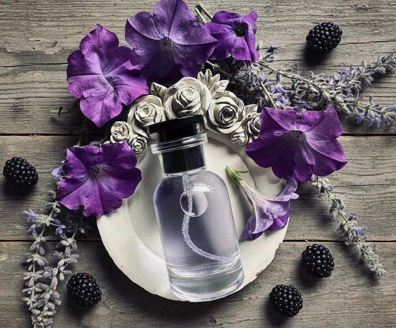 May include: A clear glass perfume bottle with a black cap sits on a white circular plate. The bottle is surrounded by vibrant purple petunia flowers, sprigs of lavender, and several dark blackberries, creating a rich and textured arrangement.