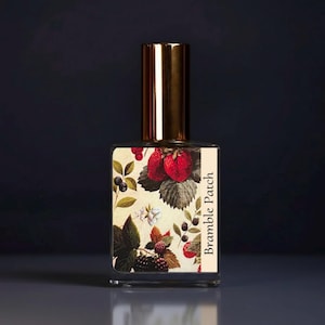 May include: A rectangular glass perfume bottle with a metallic gold cap. The label features a botanical illustration of red strawberries, dark blackberries, and green leaves on a cream background with the text Bramble Patch printed vertically.