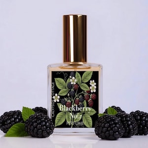 May include: A clear glass perfume bottle with a gold cap and a rectangular label featuring a botanical illustration of blackberries, green leaves, and white flowers. The label text reads Calypsos Cottage Blackberry Noir Perfume.