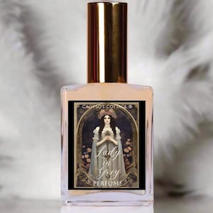 May include: A perfume bottle with a gold cap and a label featuring a woman in a grey dress and floral crown. The label reads "Lady in Grey PERFUME" and "CALYPSO'S COTTAGE ®". The bottle is clear with a light peach-colored liquid.