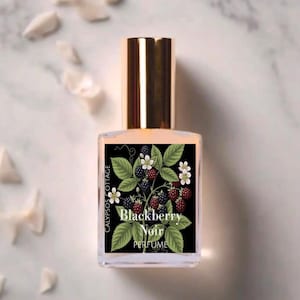 May include: A clear glass perfume bottle with a gold cap featuring a black rectangular label. The label displays a botanical illustration of green leaves, white flowers, and dark blackberries with the text Calypsos Cottage Blackberry Noir.