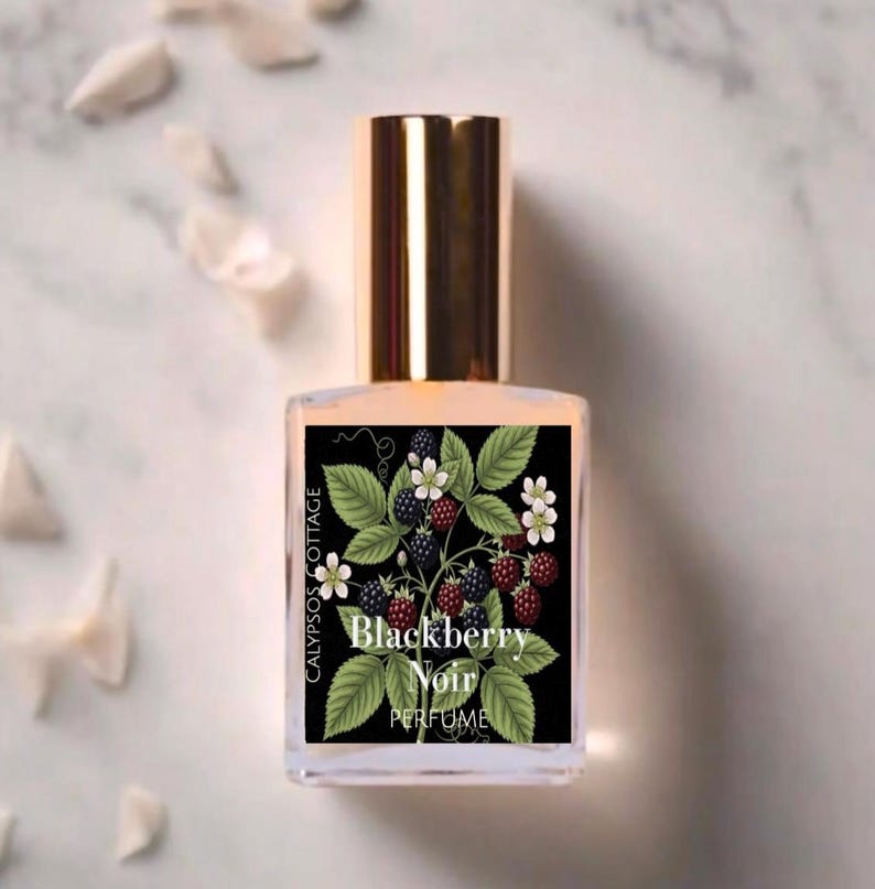 May include: A clear glass perfume bottle with a gold cap featuring a black rectangular label. The label displays a botanical illustration of green leaves, white flowers, and dark blackberries with the text Calypsos Cottage Blackberry Noir.
